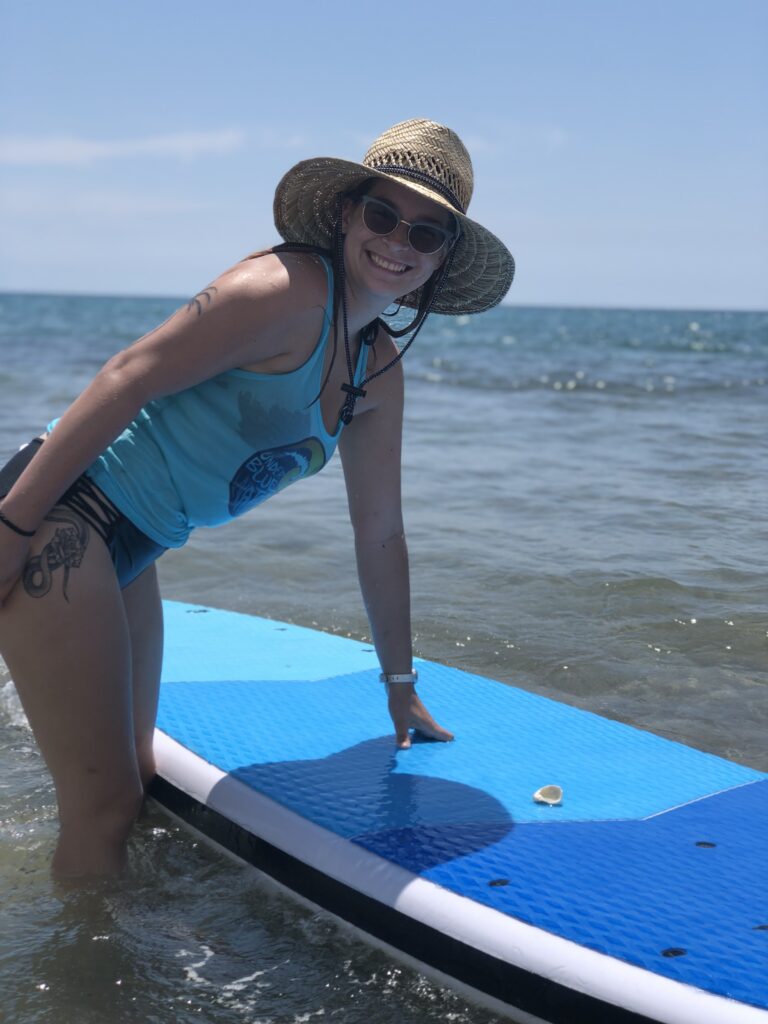 Under Blue Waters instructor smiling beside a paddleboard in the shallows